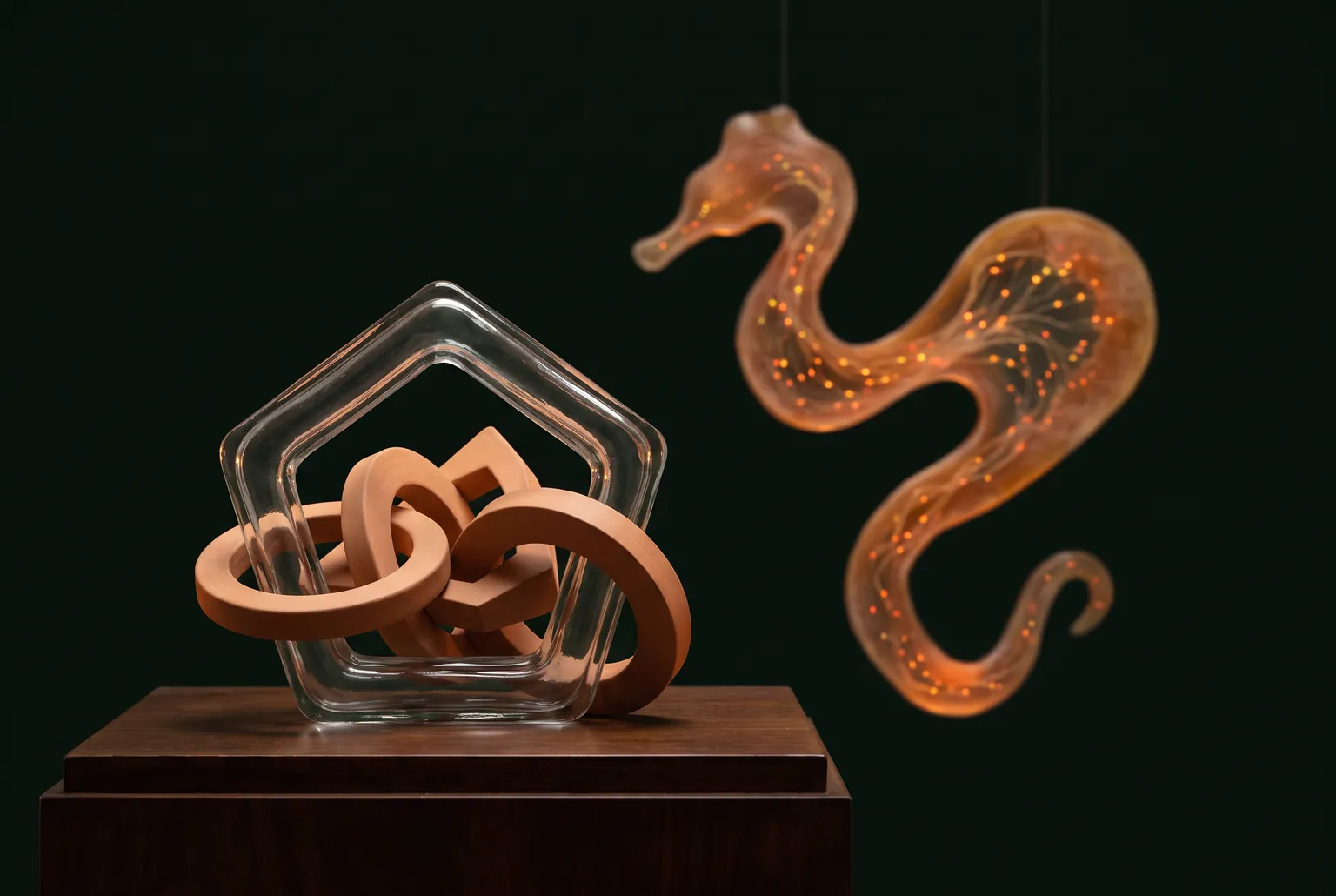 Illustrative still life of a clear glass ring entwined with terracotta ceramic rings on a wooden plinth, with a glowing seahorse-shaped form suspended behind it.
