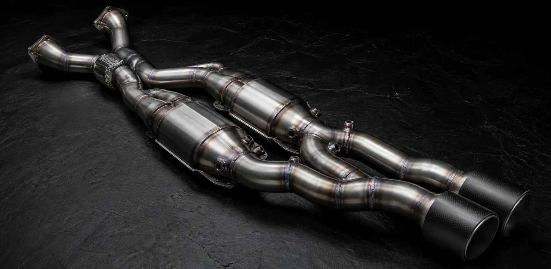 Illustrative material study of lightweight fabricated exhaust pipework and a pair of race catalytic converters on a dark work surface.