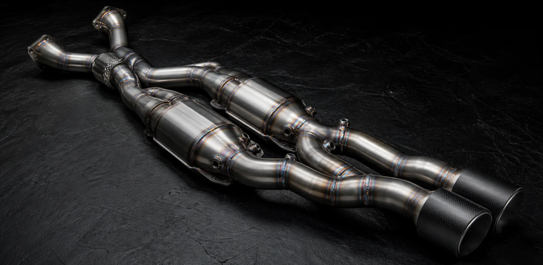 Illustrative material study of lightweight fabricated exhaust pipework and a pair of race catalytic converters on a dark work surface.